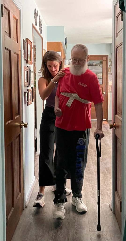 Dr. Jenna Smith walking beside a client in his own hallway, steadying him with a gait belt as he uses a cane
