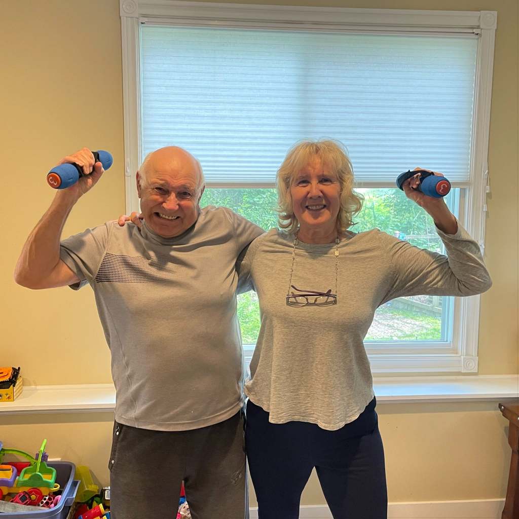 An older couple at home, grinning, each raising a hand weight